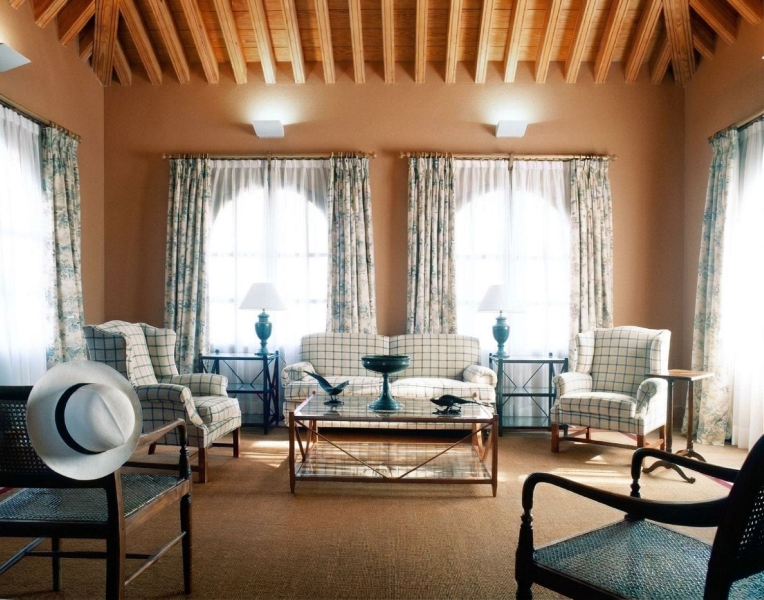 A cozy living room with a high wooden ceiling, checkered armchairs, a central coffee table, and two lamps on side tables.
