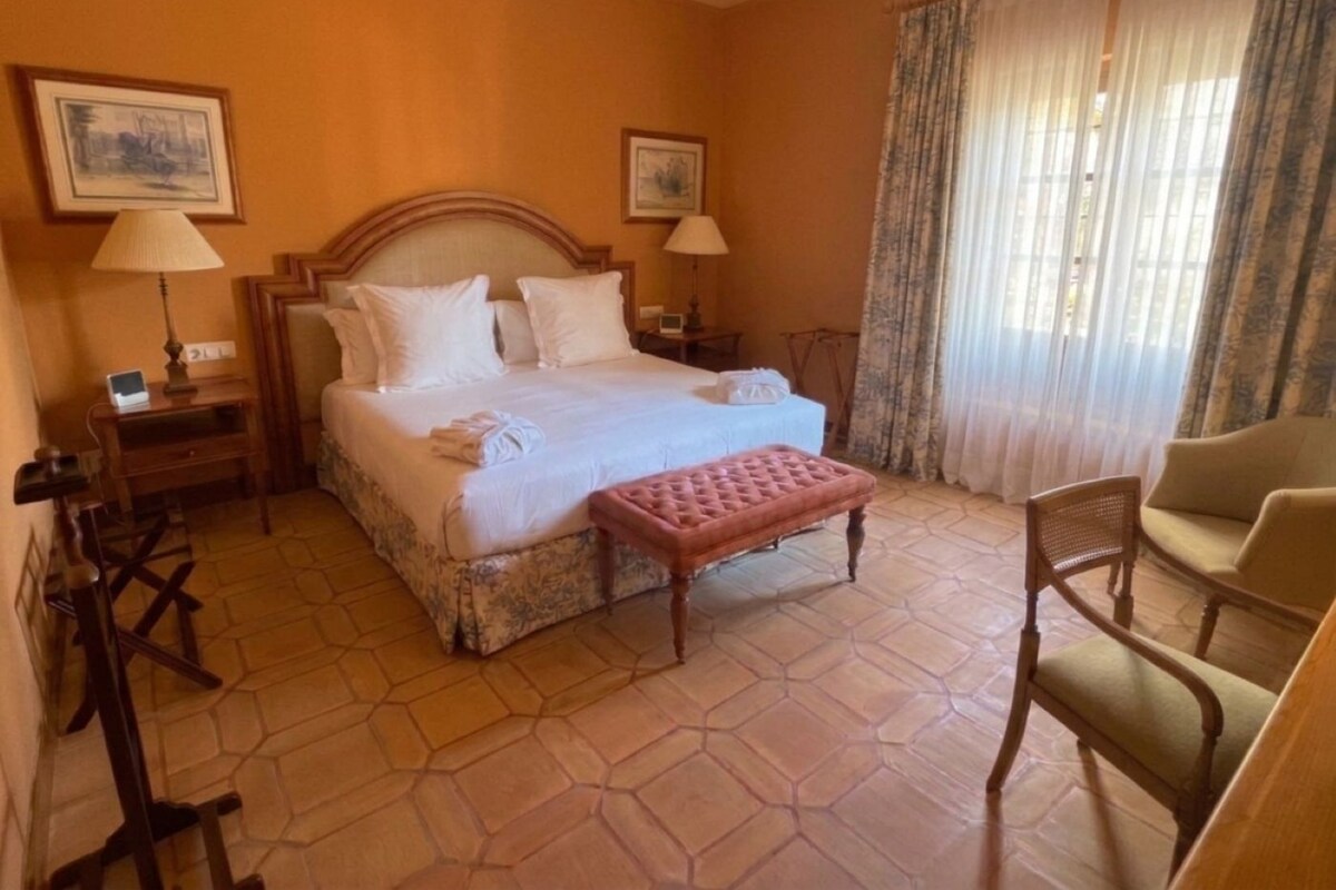 Una habitación de hotel con una cama grande, dos mesitas de noche, una silla y una ventana con cortinas.