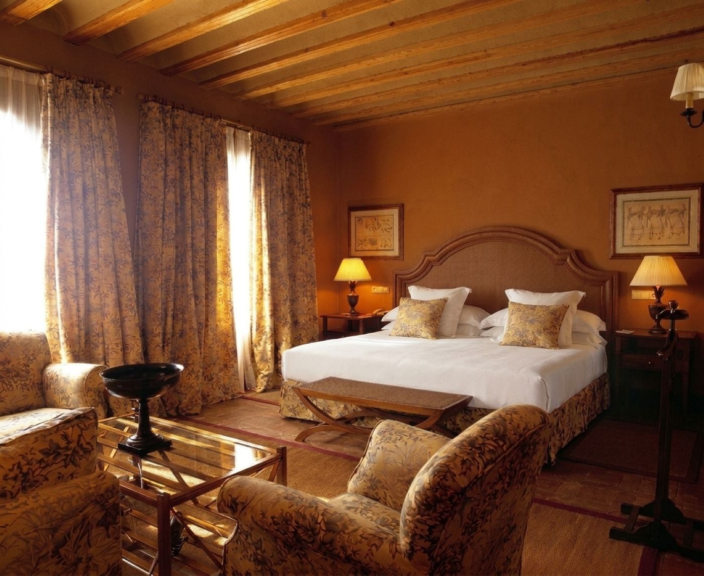 A hotel room with a large bed, two bedside tables with lamps, a wooden coffee table, floral-patterned armchairs, and curtains.