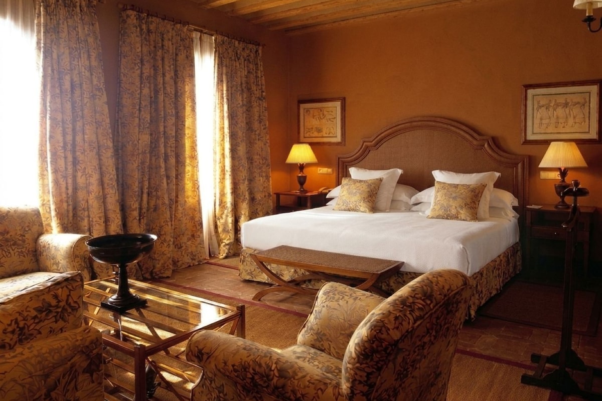 Una habitación de hotel con una cama grande, dos mesitas de noche con lámparas, una mesa de centro de madera, sillones con estampado floral y cortinas.