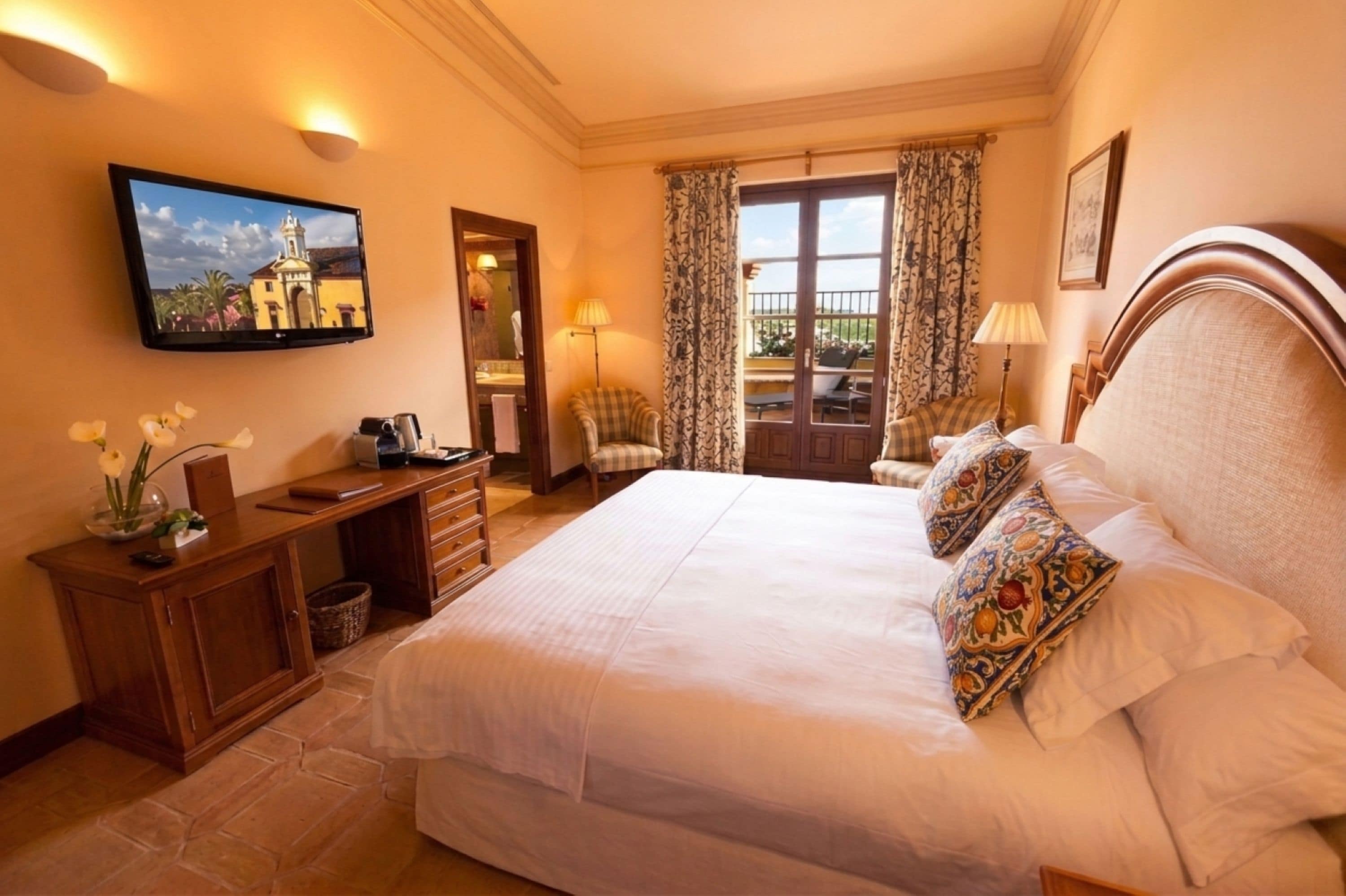 A hotel room with a bed, a desk, a chair, a TV, and a balcony with curtains.