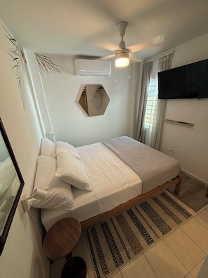 Ein Schlafzimmer mit einem Bett, einem Deckenventilator, einem an der Wand montierten Fernseher und einem Fenster mit Vorhängen.