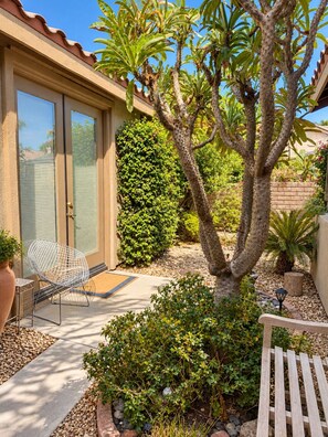 A patio with a white chair, a small table, a tree, and a hedge. - Redford Casita, 1BD/1BA, Across from Empire Polo, King Bed (Indio)