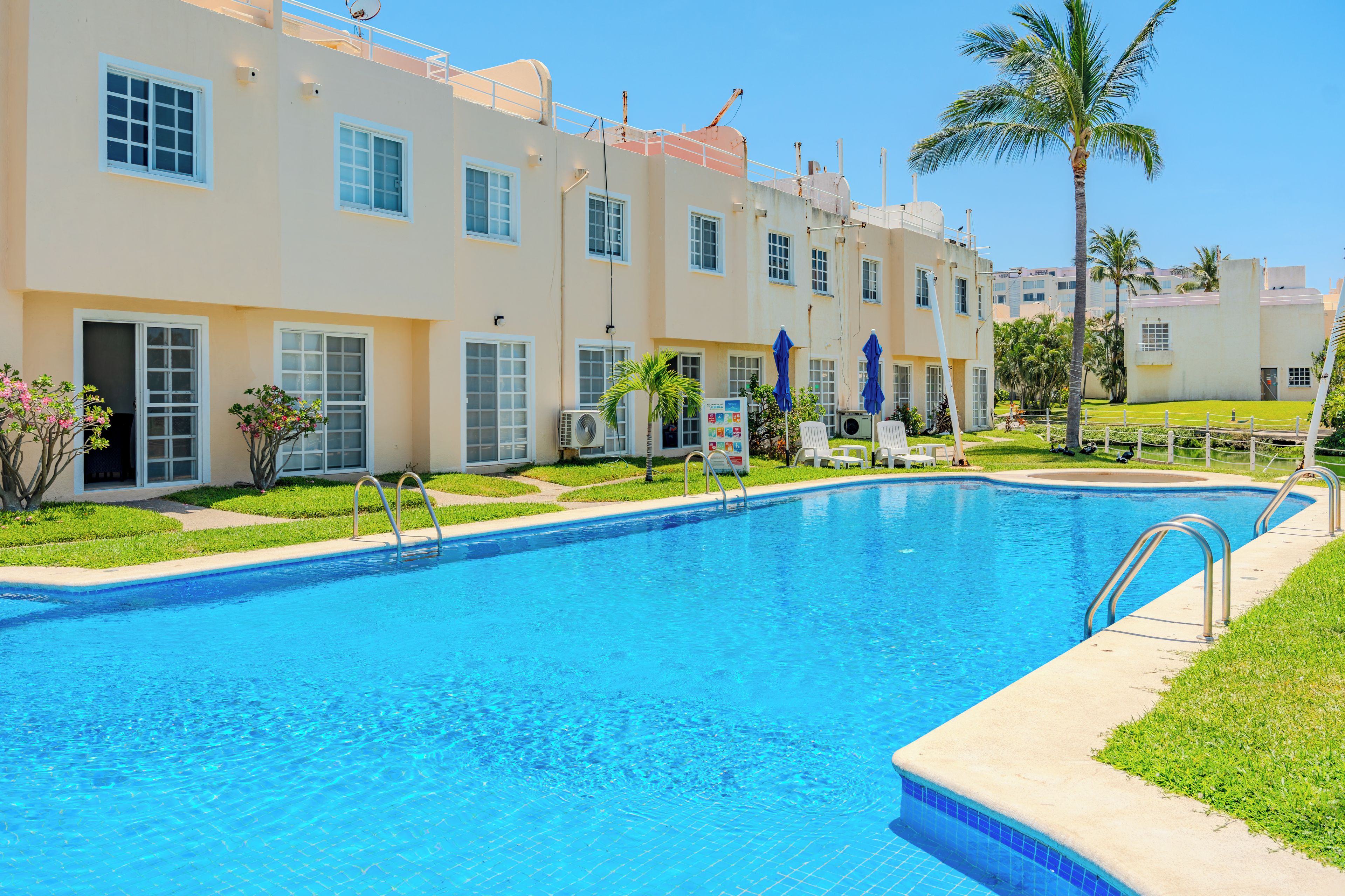 Una piscina con borde de baldosas, rodeada de césped y un edificio con varias ventanas.
