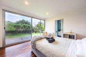 A bedroom with a large sliding glass door leading to a deck, a bed with a blanket, a bedside table with books, and a nightstand.