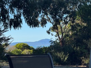 A view of a natural landscape with a chair in the foreground, trees, and a distant view of the sea and mountains.