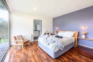 A modern bedroom with a large bed, a wooden chair, a small bedside table, and a view through a glass door. - Proms Edge - Saltbush Villa (Yanakie)