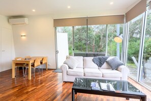 A modern living room with a glass coffee table, a white sofa with cushions, a dining table with chairs, and a large window offering a view of greenery. - Proms Edge - Saltbush Villa (Yanakie)