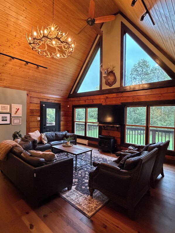 A spacious living room with a high ceiling, a chandelier, a fireplace, and large windows offering a view of the outdoors.