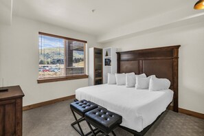 A bedroom with a large bed, a wooden headboard, a window with blinds, a bedside table, and a bench. - Silverado 310 | Canyons Village | Pool, Hot Tub & Walk to Ski (Park City)