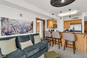 Sala de estar com sofá, duas poltronas, mesa de centro e área de cozinha com armários de madeira e geladeira.