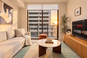Una sala de estar moderna con un sofá, una mesa de centro, un televisor mostrando Netflix y un ventanal con vista a un edificio.