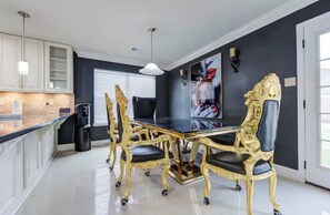 Un espace repas avec une table et des chaises noires et dorées, un plafond blanc et une grande fenêtre avec des stores.