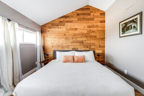 Ein Schlafzimmer mit schrägem Dach, Holzpaneelen, einem Bett mit weißer Bettwäsche und einem Nachttisch mit Lampe.