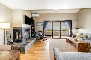 A living room with a fireplace, a sofa, a coffee table, and a view of the outdoors. - New! Ski-In/Ski-Out Condo w/ Mountain Views, Fireplace & Balcony (Whitefish)