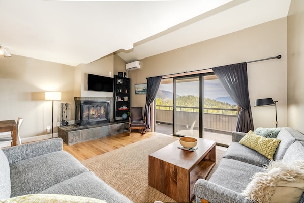A spacious living room with a fireplace, a sofa set, a coffee table, and a view of the outdoors through a sliding glass door. - New! Ski-In/Ski-Out Condo w/ Mountain Views, Fireplace & Balcony (Whitefish)