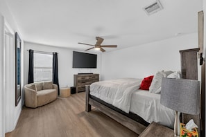 Une chambre à coucher moderne avec un lit, un ventilateur de plafond, un téléviseur, une commode en bois et une fenêtre avec des stores.