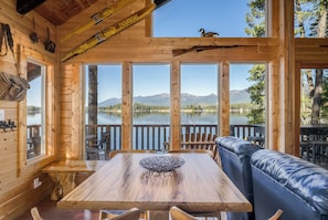Une cabane en bois comprenant un coin repas, un canapé et offrant une vue sur un lac et des montagnes.