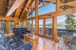 A wooden cabin interior with a large window, a leather sofa, and a dining table. - New! Arrive by Boat | Lakefront Island Cabin on Echo Lake w/ Dock & Views (Bigfork)