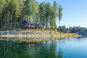 A lakeside cabin with a deck and a stone wall, surrounded by tall trees. - New! Arrive by Boat | Lakefront Island Cabin on Echo Lake w/ Dock & Views (Bigfork)