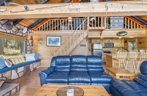 A cozy cabin interior with a leather sofa, wooden dining table, and a kitchen area. - New! Arrive by Boat | Lakefront Island Cabin on Echo Lake w/ Dock & Views (Bigfork)