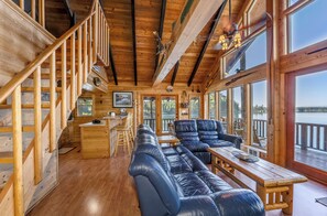 A cabin interior with a wooden staircase, a dining area, a sofa, and large windows overlooking a body of water. - New! Arrive by Boat | Lakefront Island Cabin on Echo Lake w/ Dock & Views (Bigfork)