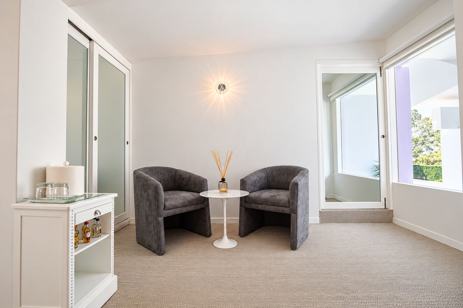 A modern room with two grey armchairs, a small round table, a glass cabinet with bottles, and a wall-mounted light fixture.