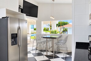 A modern kitchen with a stainless steel refrigerator, a flat-screen TV mounted above the counter, and a dining area with a glass table and metal chairs.