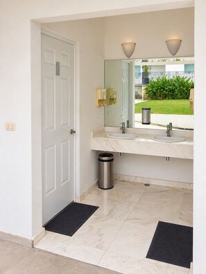 A bathroom with a white door, two sinks, a mirror, and a trash can.