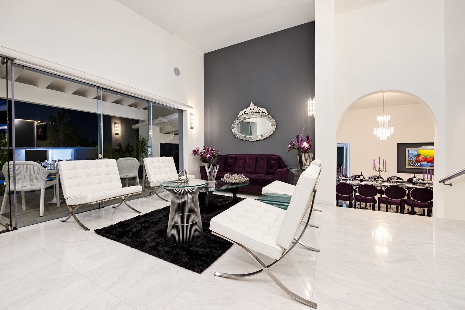 A modern interior with a dining area featuring white upholstered chairs, a glass-top table, and a dark rug.