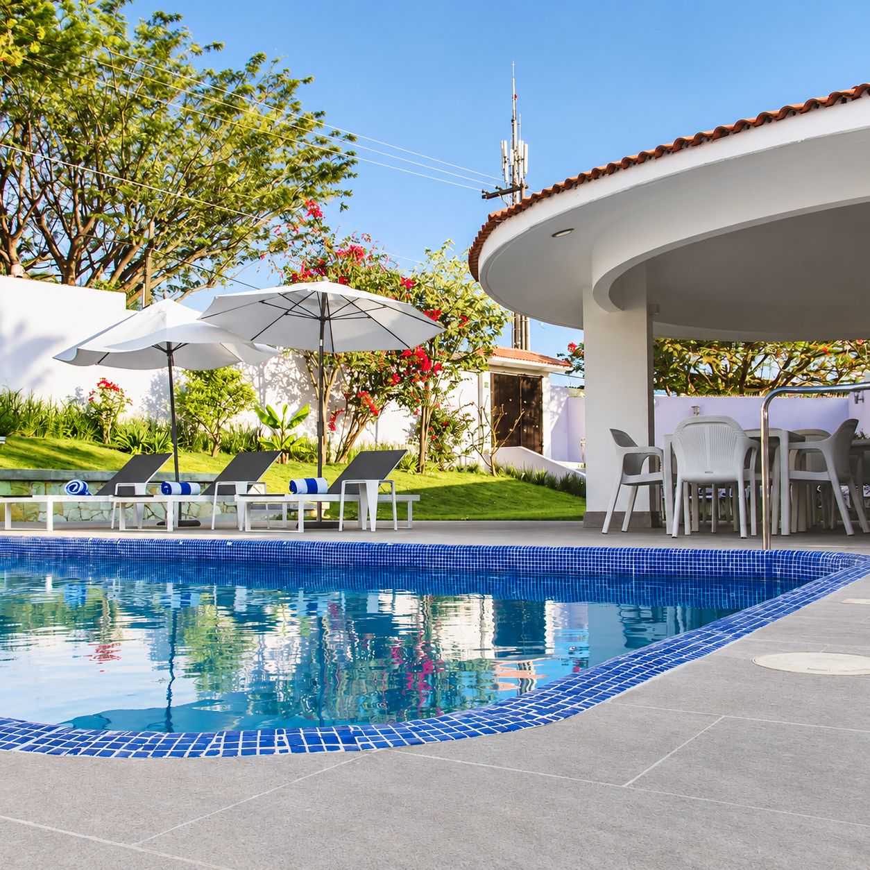 A swimming pool with lounge chairs and umbrellas, surrounded by greenery and a building with a tiled roof.