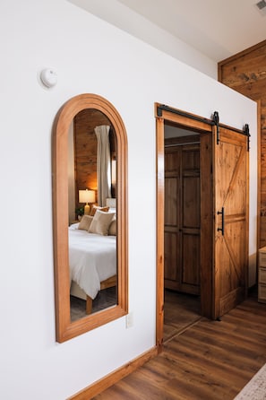 Een slaapkamer met een spiegel met houten lijst, een bed met wit beddengoed en een houten garderobe met schuifdeur.
