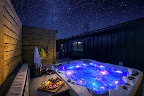 Un bain à remous sous un ciel étoilé, entouré de murs en bois, avec un plateau de nourriture et des verres de vin.