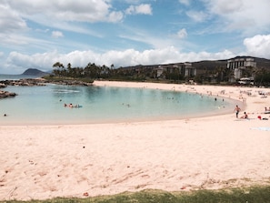 A beach with clear water, a sandy shore, and a resort building in the background. - Kai Lani – Beautiful Ocean View Condo In Paradise (Kapolei)