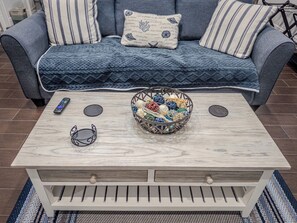 Ein Wohnzimmer mit einer Couch, einem Couchtisch, einer Schüssel voller bunter Kugeln, einer Fernbedienung und einer Sonnenbrille.