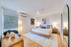 Een slaapkamer met een bed, bedlampjes, een wandairconditioner, een raam met zonwering en een spiegel.