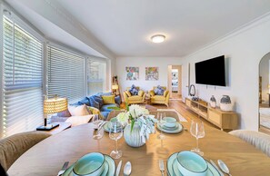 Ein helles Wohnzimmer mit Essbereich, gedeckt für eine Mahlzeit, ausgestattet mit einer Couch, einem Couchtisch und einem an der Wand befestigten Flachbildfernseher.