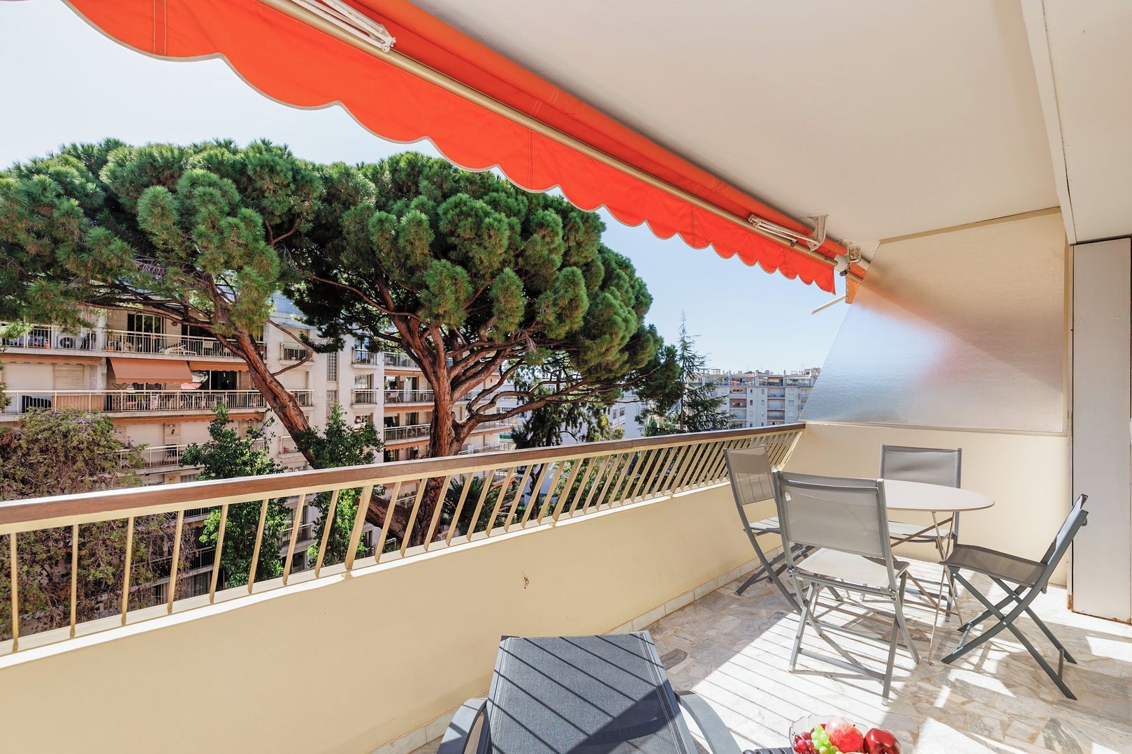 A balcony with a table and chairs, a red awning, and a view of a tree and buildings.