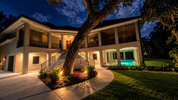 A two-story house with a swimming pool, a large tree in the front yard, and a well-lit exterior.