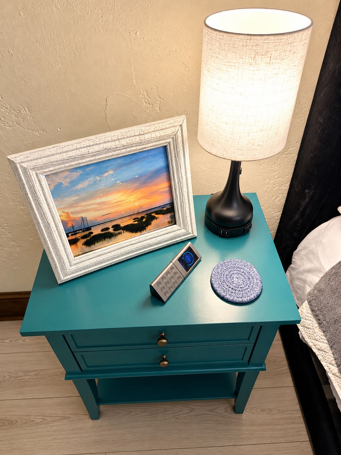 A teal bedside table with a framed picture, a remote control, and a lamp.