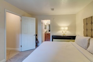 Habitación de hotel con cama, mesita de noche con lámpara, una puerta y vista al pasillo.