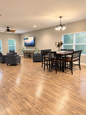 A spacious living room with a wooden floor, a dining area with a table and chairs, a flat-screen TV mounted on the wall, and a ceiling fan. - 3-bedroom house in amazing Fort Walton Beach within 6 minutes from the beach. (Fort Walton Beach)