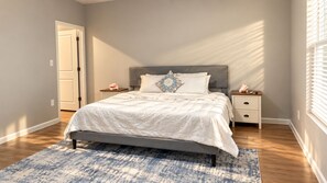 Une chambre à coucher avec un lit, des tables de chevet, une commode, une porte et une fenêtre avec des stores.