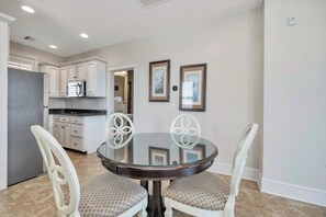 A dining area with a round glass table and white chairs, a kitchen with white cabinets and a refrigerator, and two framed pictures on the wall. - One bedroom with great ocean views! (St. Simons Island)