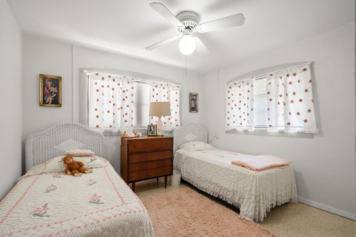 A bedroom with two single beds, a dresser, a ceiling fan, and two windows with floral curtains.