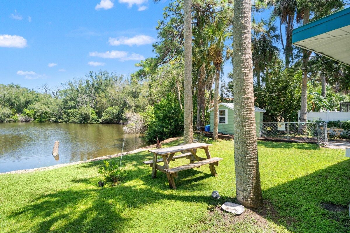 A picnic table by a lake with a small shed and a house in the background.
