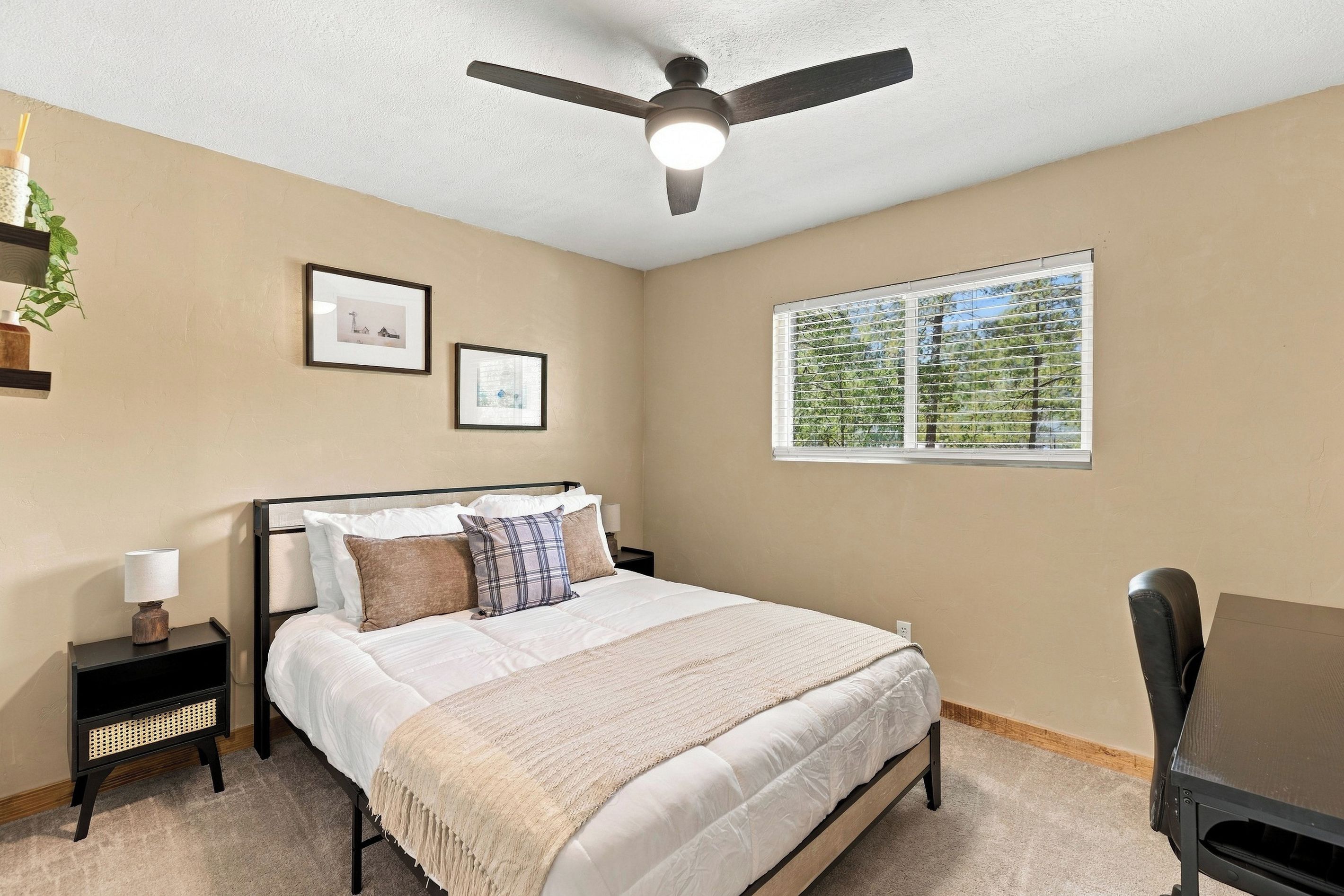 A bedroom with a bed, a ceiling fan, a window with blinds, and two framed pictures on the wall.
