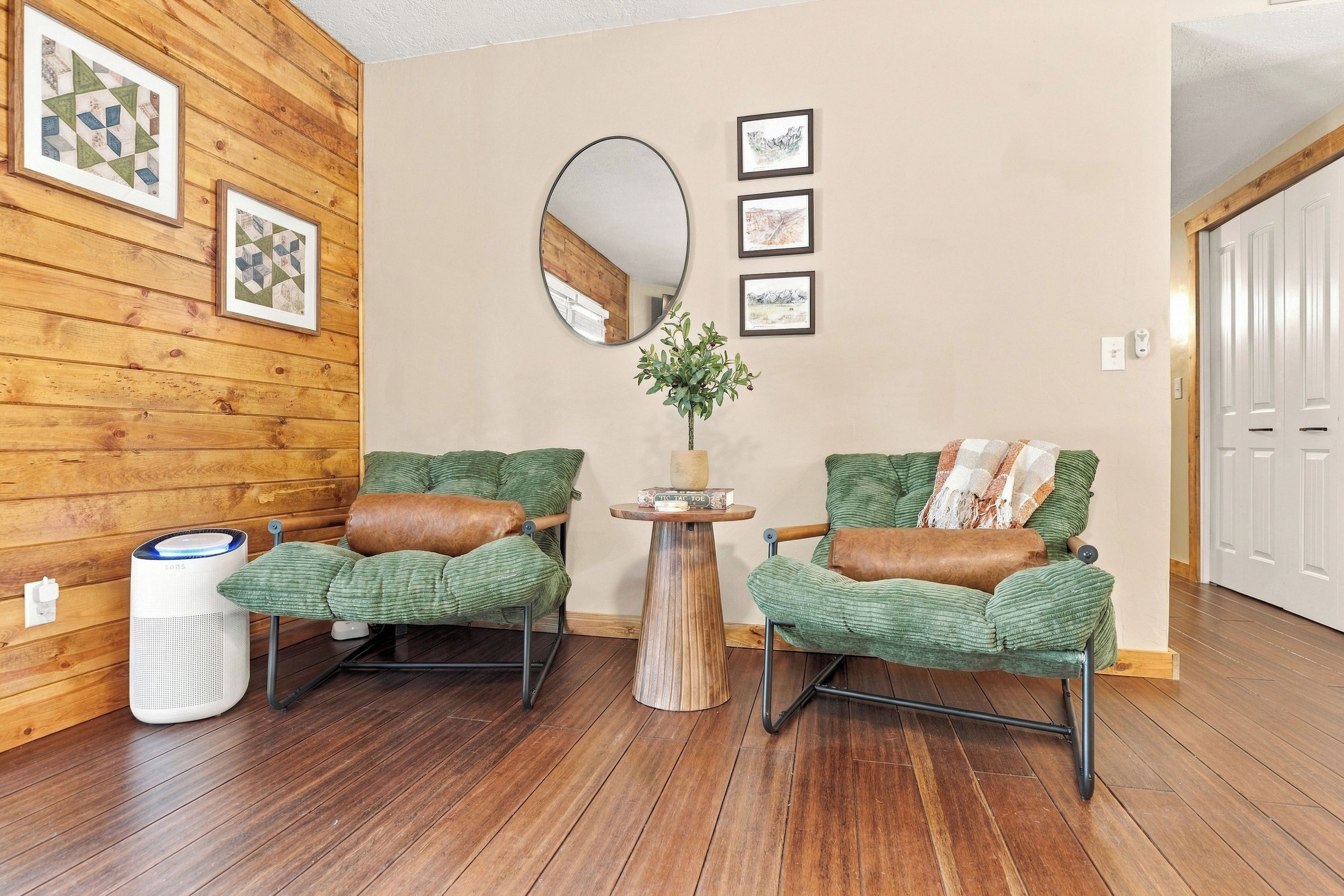 A room with wooden walls, two green upholstered chairs with brown cushions, a round mirror, and a small wooden table with a plant.