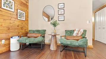 A room with wooden walls, two green upholstered chairs with brown cushions, a round mirror, and a small wooden table with a plant.