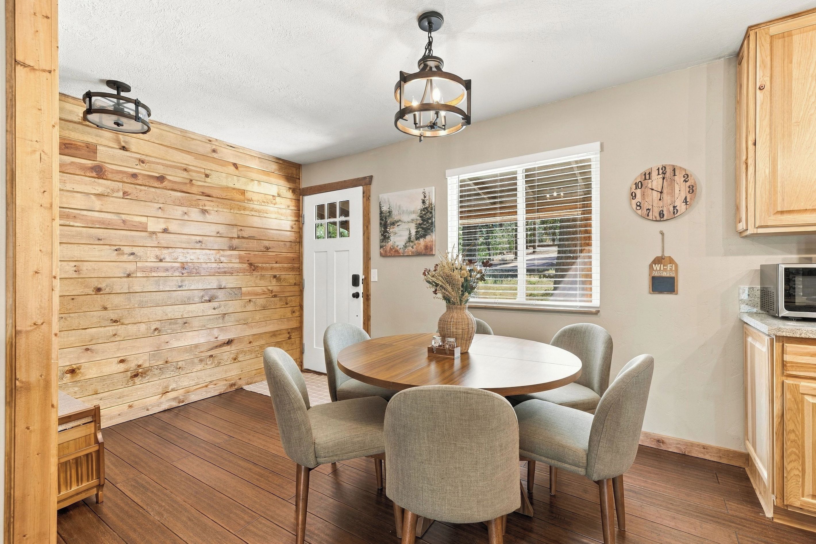 A dining area with a wooden table and chairs, a ceiling light fixture, a window with blinds, and a clock on the wall.